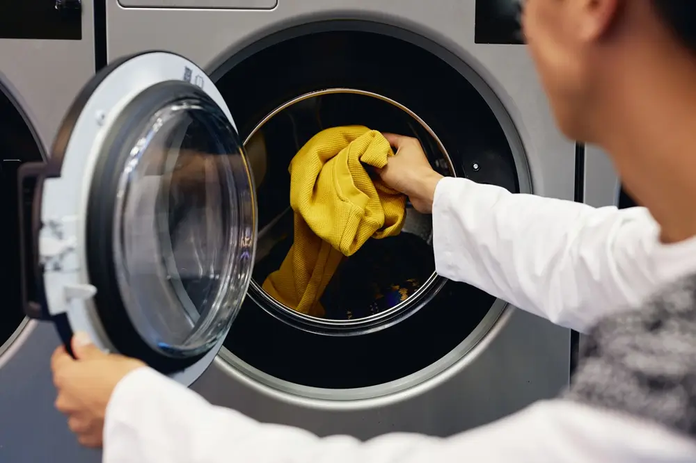 A person placing a yellow garment into a front-loading washing machine. A person placing a yellow garment into a front-loading washing machine.